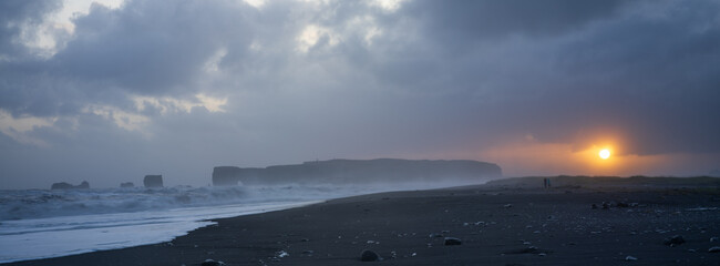 Reynisfjara black sand beach at sunset. Moddy skies and golden sun. A couple (small) walking along beach. South Coast of iceland. Panorama image.