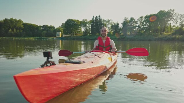 Positive man with safety equipment sitting in canoe on lake. Holding oar while looking at camera. Cheerful smiling during resting outdoors. On boat fixing action camera. Getting ready to swim.