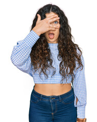 Young hispanic girl wearing casual clothes peeking in shock covering face and eyes with hand, looking through fingers with embarrassed expression.