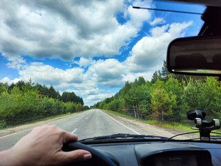 view through car windshield of natural landscape with road, green tree, white clouds. Hand of woman on the steering wheel of car. Female traveler driving on trip or journey in spring or summer