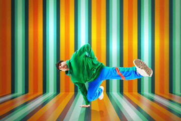 Young flexible sportive man dancing breakdance in green and blue sport uniform outfit against colorful striped background.