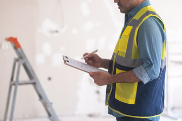 Focused engineer editing plan of apartment renovation standing against ladder near shabby wall worker in professional uniform holding clipboard and pen in hands