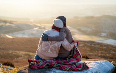 Back view of the happy couple in love sitting on top of a mountain enjoying a sunset winter...