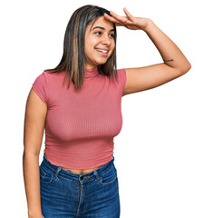 Young hispanic girl wearing casual t shirt very happy and smiling looking far away with hand over head. searching concept.