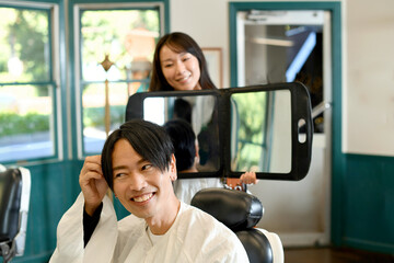 男性客に鏡を見せる美容師のアジア人女性 - obrazy, fototapety, plakaty