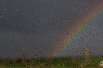 Focus on  rain drops on a window. In the distance intentionally out of focus, very dark storm clouds and a rainbow. 