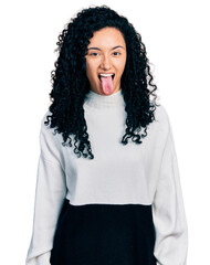 Young hispanic woman with curly hair wearing casual sweater sticking tongue out happy with funny expression. emotion concept.