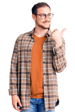 Young handsome caucasian man wearing casual clothes and glasses smiling with happy face looking and pointing to the side with thumb up.