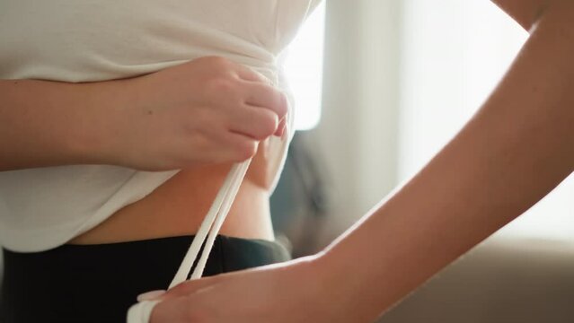 Woman adjusts tank top with ropes in sunny room closeup. Slim lady wearing white shirt with ties ribbons on side at home slow motion. Stylish clothes