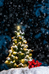 A glowing Christmas Tree with star shaped lights outside at night while it is snowing with red baubles and bows sitting in the snow