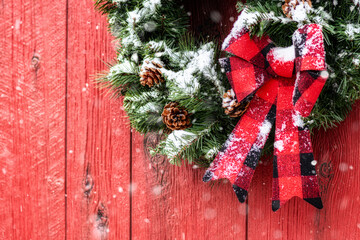 A Christmas wreath made from evergreens, pine cones and a red and black plaid bow hanging on a red barn while it is snowing. © CMANNPHOTO