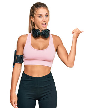 Young blonde woman wearing gym clothes and using headphones smiling with happy face looking and pointing to the side with thumb up.