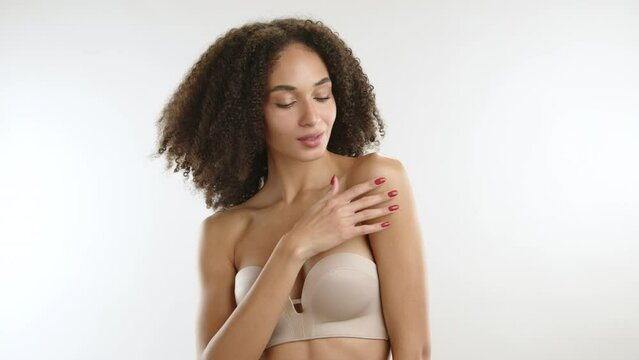 Curly-haired thin build sexy beauty wearing nude strapless bra running her fingers with red manicure on perfect body standing on white background. High quality 4k footage
