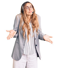 Young beautiful blonde woman wearing call center agent headset clueless and confused expression...