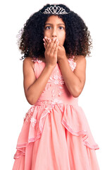 African american child with curly hair wearing princess crown shocked covering mouth with hands for...