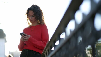 Woman looking at cellphone device outside in city street leaning on metal rail in daylight. 20s female person texting message on phone, using modern technology