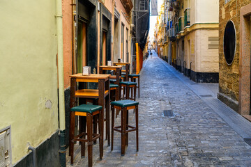 A sidewalk cafe with empty tables and stools along a narrow pedestrian cobblestone alley in the...