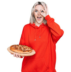 Young blonde girl holding italian pizza smiling happy doing ok sign with hand on eye looking...