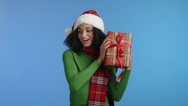 Smiling attractive young woman with dark short hair with red checkered scarf holding box wrapped in checkered paper, shaking xmas gift, posing for camera. High quality 4k footage