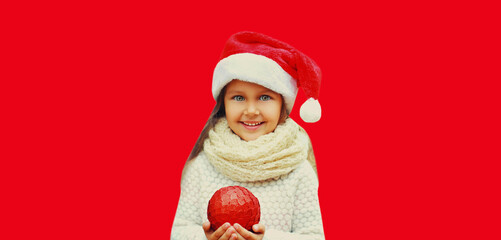 Christmas portrait of happy smiling little girl child in red santa hat with ball toy isolated on studio background