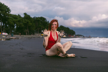 Happy woman tourist in red bodysuit shows dirty hands. Sitting on the black sand near the water, waves on the sea. Rain clouds on the horizon.