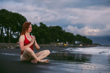 Happy woman tourist in red bodysuit Sitting in the lotus position, yoga on black sand, stormy waves...