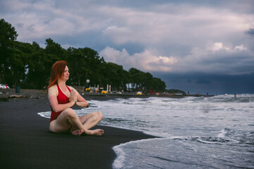 Happy woman tourist in red bodysuit Sitting in the lotus position, yoga on black sand, stormy waves on the sea. Danger for swimming. Rain clouds on the horizon. Copy space. Ureki besch, Georgia