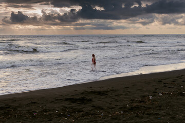 A woman walks into stormy waves dangerous for swimming, view in the distance. Standing on black sand, big stormy waves on the sea. Danger. Rain clouds on the horizon.