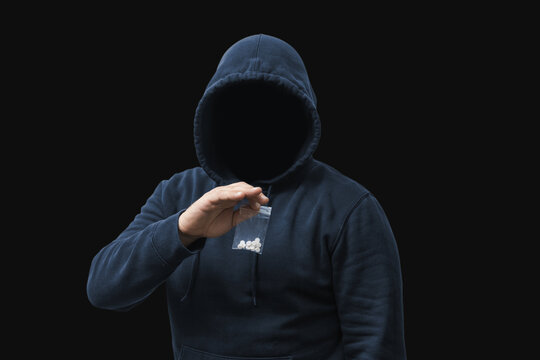 Faceless man in a hood holds transparent plastic bag with white pills hard drugs on dark background, anonymous drug dealer or gangster sells narcotics