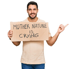 Young hispanic man holding mother nature is crying protest cardboard banner smiling happy pointing with hand and finger to the side