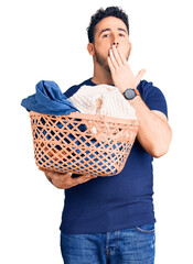 Young hispanic man holding laundry basket covering mouth with hand, shocked and afraid for mistake....