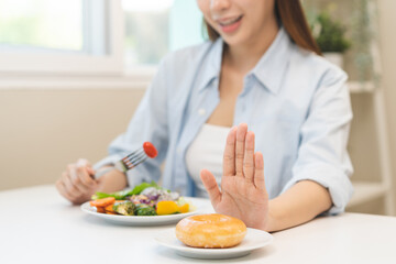 Diet concept, close up young woman, girl using hand push out, stop sweet donut, dessert or junk food on plate, choose green vegetables salad, eat low fat for good health. Female getting weight loss.