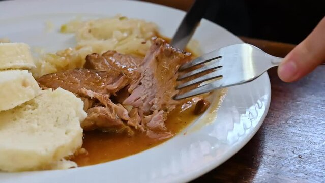 Prague, Czech Republic, August 4,2023. Close-up in slow motion with one of the typical dishes: stewed pork with sauerkraut and bread for dipping. Hands with fork and knife cut a piece of meat.