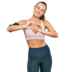 Beautiful young blonde woman wearing sportswear and arm band smiling in love showing heart symbol and shape with hands. romantic concept.