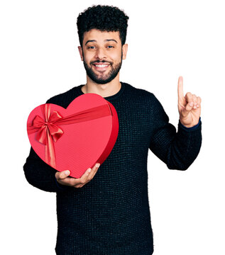 Young arab man with beard holding valentine gift smiling with an idea or question pointing finger with happy face, number one