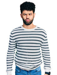 Young arab man with beard wearing casual striped sweater making fish face with lips, crazy and comical gesture. funny expression.