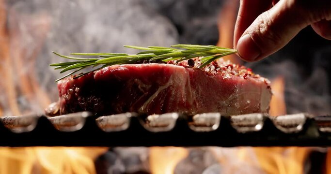 Close-up. Hand gently places sprig of fragrant rosemary onjuicy steak sprinkled with seasonings. Flickering fire of barbecue sets stage for unforgettable summer party. Flickering flames in background.