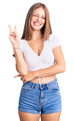 Young beautiful blonde woman wearing casual white tshirt smiling with happy face winking at the camera doing victory sign with fingers. number two.