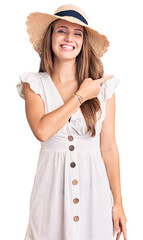 Young beautiful blonde woman wearing summer dress and hat cheerful with a smile of face pointing with hand and finger up to the side with happy and natural expression on face