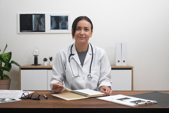 Smiling female doctor looks at camera sitting at table in office with x-rays pictures on wall young therapist reviews notes positive medic waits for patients in clinic