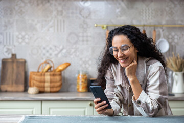 A young beautiful woman at home in kitchen smiles contentedly, browses social networks, online...
