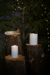 White LED candles that stand on fir tree stumps under fir trees with green needles in which Christmas lights are installed that create a bokeh effect.