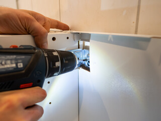 Mounting a corpus furniture part on the wall. Fixing the kitchen that nothing can be moved away. Using the electric drilling machine to insert a screw into the wall of the apartment.