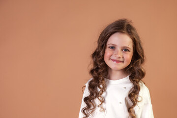 Lovely smiling child model in white wear at beige background, looking at camera. Portrait of kid cover girl 7 year old with curly hair, studio shot. Children, childhood concept. Copy ad text space