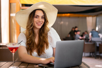 Stylish woman in big straw hat working remotely from a cafe, drinking cocktail and using laptop,...