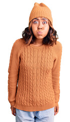 Young beautiful mixed race woman wearing wool sweater and winter hat puffing cheeks with funny face. mouth inflated with air, crazy expression.