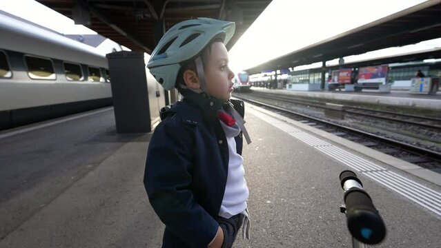 One pensive young boy standing at train platform wearing helmet staring blankly into the horizon daydreaming. Thoughtful Child with hands in pocket waiting for transportation to arrive