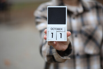 Woman in warm shirt holds calendar with date October 11 outdoors in autumn park