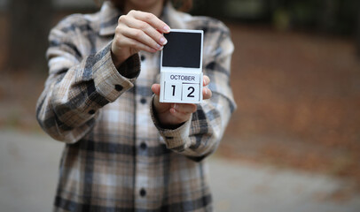 Woman in warm shirt holds calendar with date October 12 outdoors in autumn park