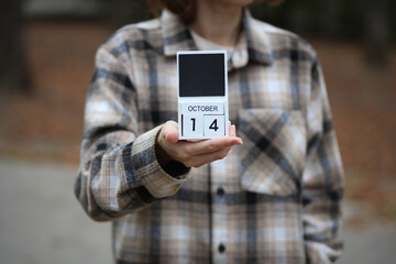 Woman in warm shirt holds calendar with date October 14 outdoors in autumn park